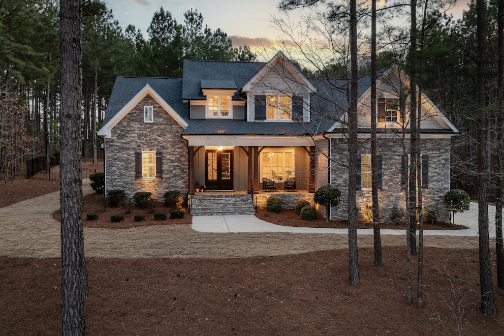 Charming modern stone house nestled among tall pine trees at dusk, emitting a warm glow.