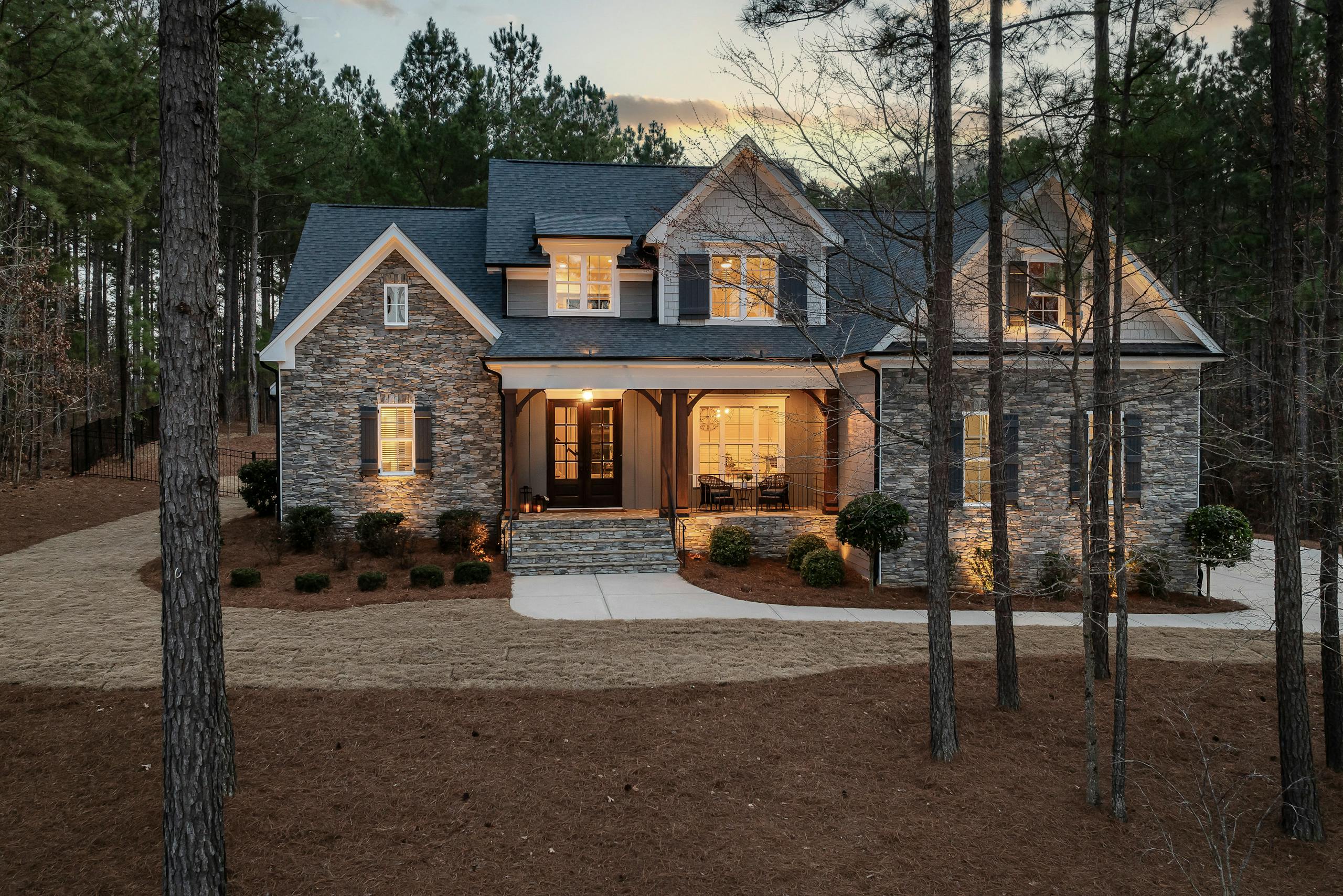 Charming modern stone house nestled among tall pine trees at dusk, emitting a warm glow.